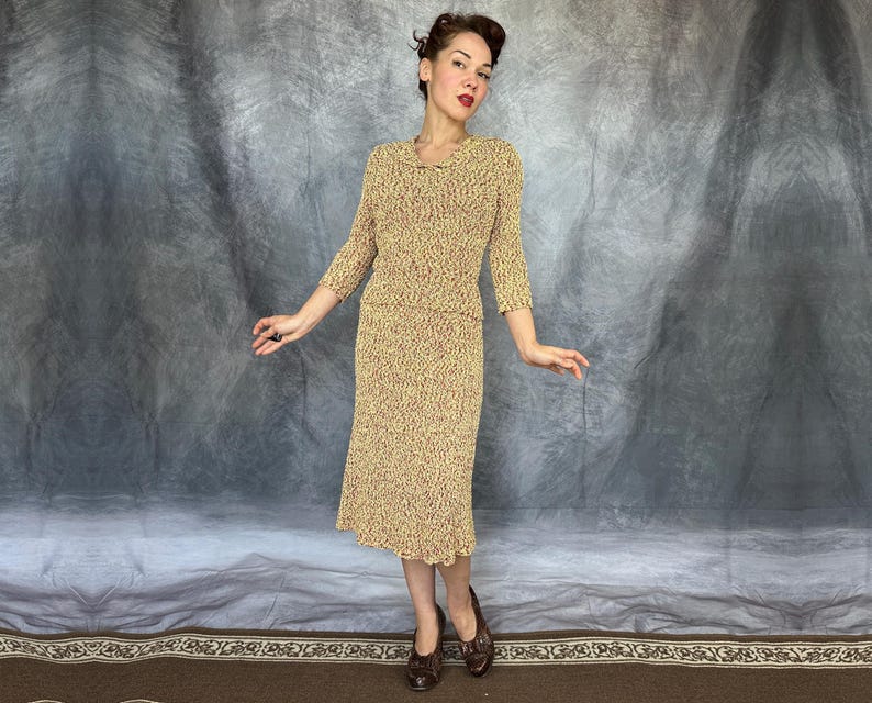 May include: A vintage two-piece dress set in a textured knit fabric. The set includes a long-sleeved top and a knee-length skirt, both in a beige color with a subtle pattern of red and brown flecks. The dress has a scalloped hem.