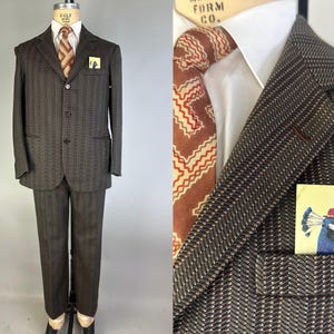 May include: A dark brown suit with a subtle herringbone pattern, featuring a three-button jacket, a white collared shirt, and a brown tie with a geometric zigzag print. A pocket square displays a colorful illustration of a peacock wearing a hat.
