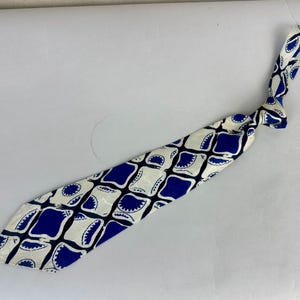 May include: A necktie featuring a repeating geometric pattern of dark blue squares with rounded corners and white shapes with scalloped edges, all separated by thin black lines. The tie is knotted at the top and displays a pointed bottom end.