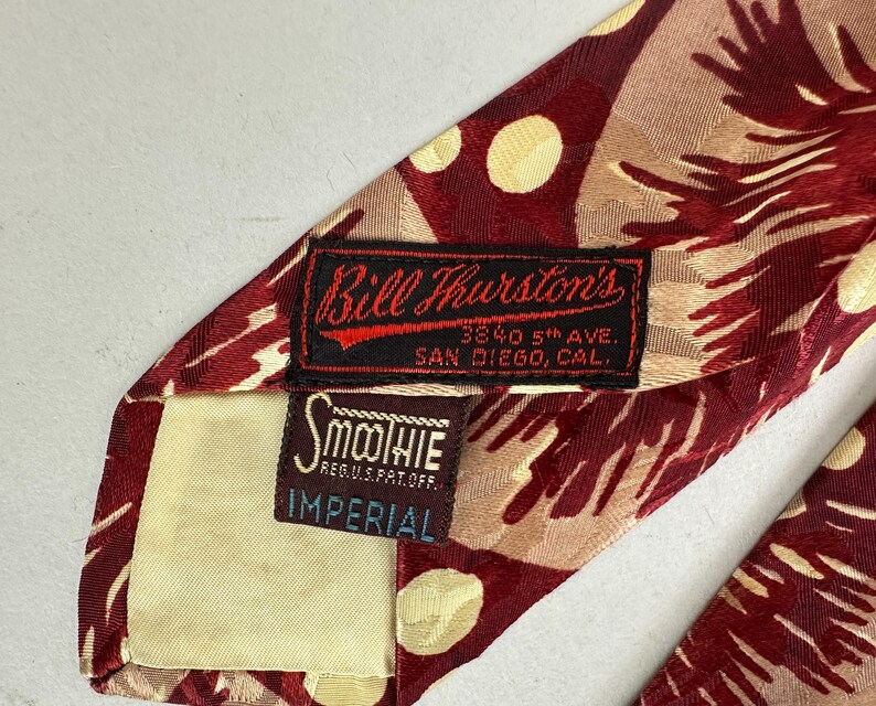 May include: A patterned necktie in burgundy and tan, with cream-colored polka dots. The label reads "Bill Thurston's, San Diego, Cal." and "Smoothie Imperial". A vintage accessory.
