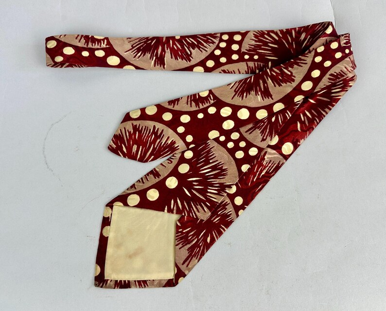 May include: A maroon tie featuring a repeating pattern of beige half-circles and white polka dots. The tie is made of a silky material. The tie's design is a vintage style.