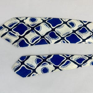 May include: A necktie featuring a repeating geometric pattern of blue and off-white shapes. The design consists of rounded blue squares alternating with off-white diamond-like forms, each containing a small circular blue detail with scalloped edges.