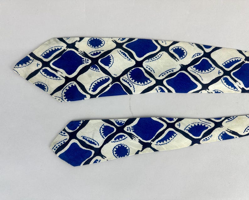 May include: A necktie featuring a repeating geometric pattern of blue and off-white shapes. The design consists of rounded blue squares alternating with off-white diamond-like forms, each containing a small circular blue detail with scalloped edges.