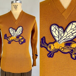 May include: A mustard yellow v-neck sweater with a purple and yellow embroidered wasp on the front. The sweater has purple trim on the sleeves and the text "WOLF MODEL FORM CO. N.Y." on the back of the collar.