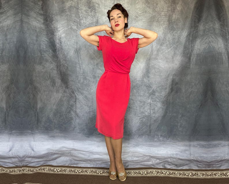 May include: A solid bright red knee-length dress featuring short sleeves, a round neckline, and a draped wrap-style bodice detail at the waist. The garment has a fitted silhouette that extends to the knees, providing a clean and simple aesthetic.