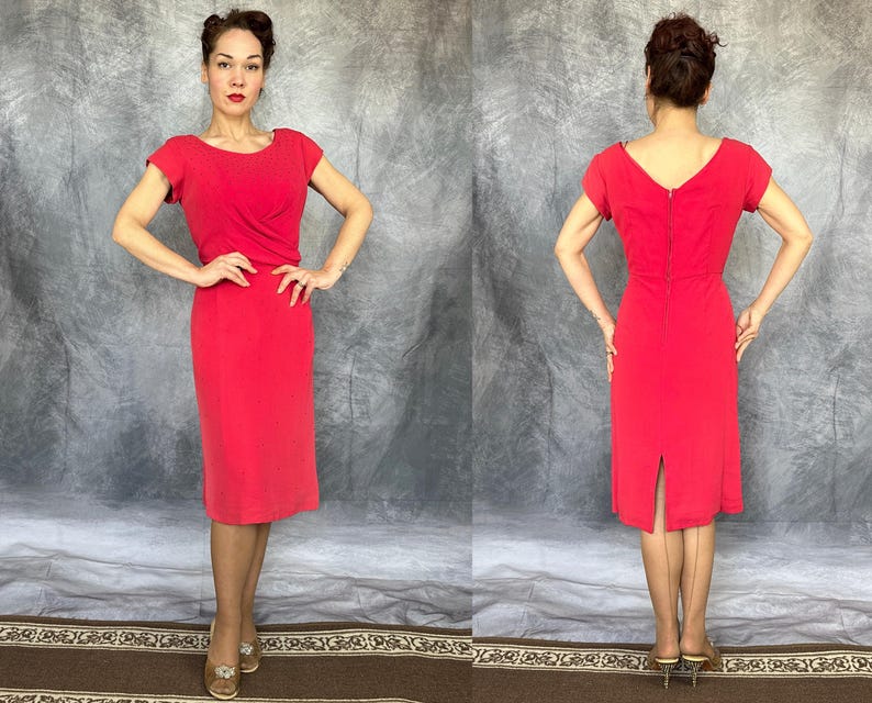 May include: A solid red knee-length dress featuring short sleeves, a boat neckline, and a fitted bodice with subtle draping at the waist. The back of the dress displays a central vertical zipper closure and a small slit at the hemline.
