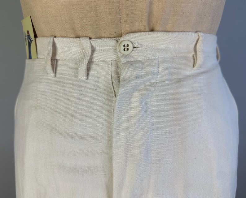 May include: Off-white trousers featuring a high-waisted design with a single button closure at the centre front. The trousers include belt loops along the waistband and a visible fly construction, presented on a neutral-toned mannequin torso.