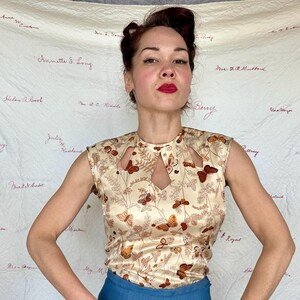 May include: A sleeveless, cream-colored top with a butterfly pattern. The top has a keyhole neckline and is paired with blue pants. The butterfly design features brown and orange butterflies and floral accents.