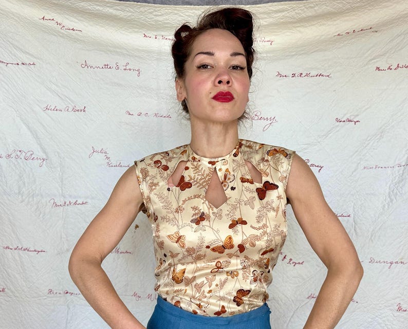 May include: A sleeveless, cream-colored top with a butterfly pattern. The top has a keyhole neckline and is paired with blue pants. The butterfly design features brown and orange butterflies and floral accents.
