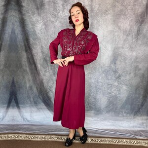May include: A burgundy dress with a long skirt and long sleeves. The dress has an embroidered bodice and a black belt. The woman is wearing black shoes.