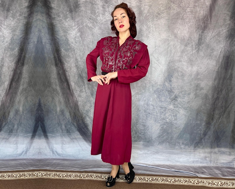 May include: A burgundy dress with a long skirt and long sleeves. The dress has an embroidered bodice and a black belt. The woman is wearing black shoes.