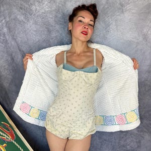 May include: A one-piece swimsuit featuring a light beige fabric with small grey polka dots, a sweetheart neckline, and thin straps. It is worn under a white, textured, open-front robe with a decorative border of pastel-colored circular embroidery.