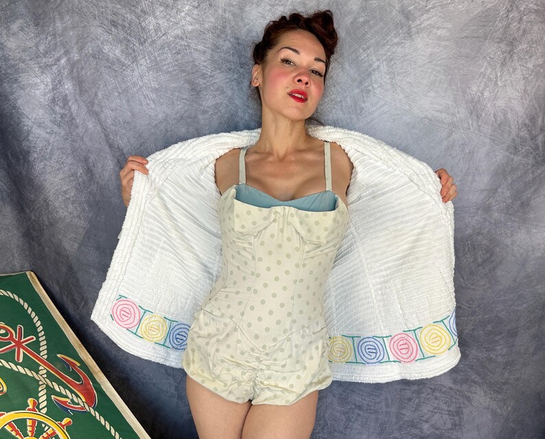 May include: A one-piece swimsuit featuring a light beige fabric with small grey polka dots, a sweetheart neckline, and thin straps. It is worn under a white, textured, open-front robe with a decorative border of pastel-colored circular embroidery.