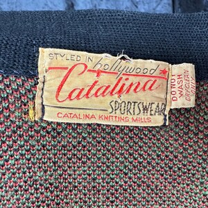May include: Close-up of a vintage Catalina Sportswear label. The label is beige with red and black text, including "Styled in Hollywood" and "Catalina Sportswear." The fabric beneath is a knit pattern in shades of green, red, and black.