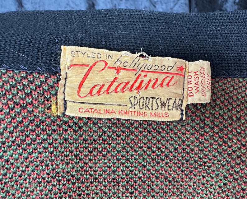 May include: Close-up of a vintage Catalina Sportswear label. The label is beige with red and black text, including "Styled in Hollywood" and "Catalina Sportswear." The fabric beneath is a knit pattern in shades of green, red, and black.