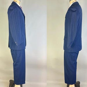 Puede incluir: Un traje azul marino con un sutil patr&oacute;n de rayas verticales. El conjunto consta de una americana de botonadura sencilla con solapas de muesca y dos botones en los pu&ntilde;os, combinada con pantalones a juego de corte recto.