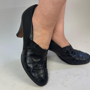 May include: Black leather pumps with a stacked heel. The shoes have a rounded toe and a pleated design on the vamp. The heels are approximately 3 inches (7.6 cm) high.