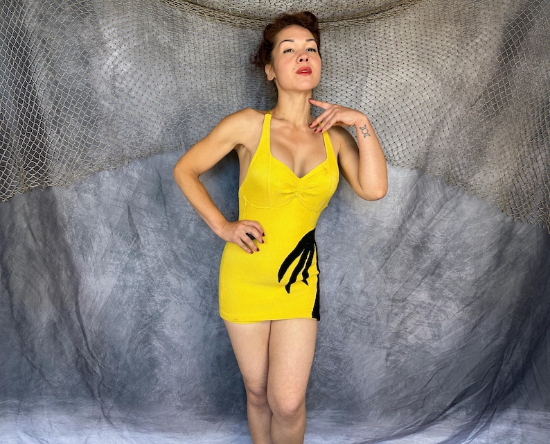 May include: A bright yellow sleeveless mini dress featuring a sweetheart neckline and a black graphic design on the lower left side. The garment has a fitted silhouette with thin shoulder straps and a solid yellow back and front bodice construction.