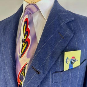 Puede incluir: Un blazer azul de rayas diplom&aacute;ticas sobre una camisa blanca de cuello y una corbata lavanda con un patr&oacute;n repetitivo de llamas amarillas, rojas y negras. Un pa&ntilde;uelo de bolsillo amarillo con un pavo real con sombrero rojo est&aacute; insertado.