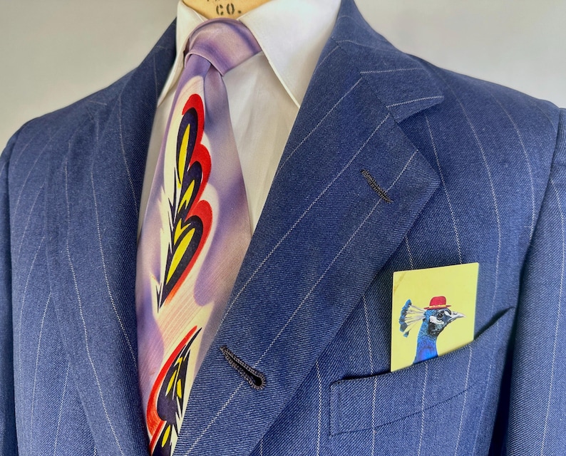 Puede incluir: Un blazer azul de rayas diplom&aacute;ticas sobre una camisa blanca de cuello y una corbata lavanda con un patr&oacute;n repetitivo de llamas amarillas, rojas y negras. Un pa&ntilde;uelo de bolsillo amarillo con un pavo real con sombrero rojo est&aacute; insertado.