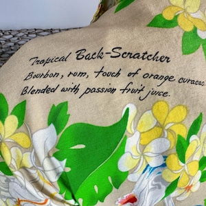 May include: Tan fabric with a tropical floral pattern and the text "Tropical Back-Scratcher" with a cocktail recipe: Bourbon, rum, orange curacao, and passion fruit juice. The design includes white, yellow, and green flowers.
