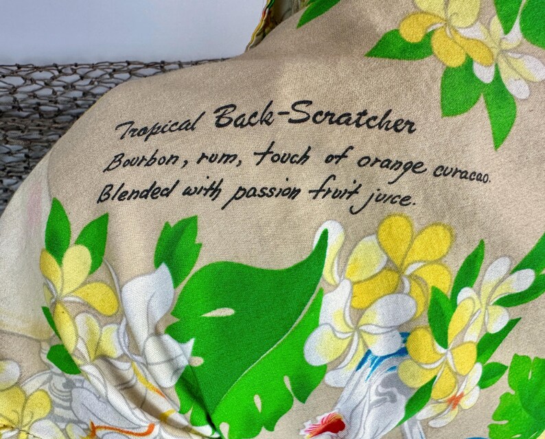 May include: Tan fabric with a tropical floral pattern and the text "Tropical Back-Scratcher" with a cocktail recipe: Bourbon, rum, orange curacao, and passion fruit juice. The design includes white, yellow, and green flowers.