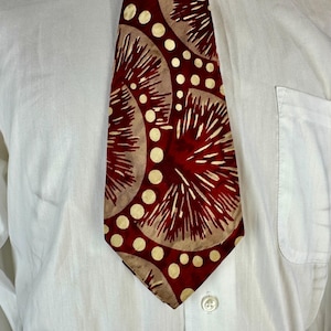 May include: A patterned tie with a red background and beige circular designs with white accents. The tie is worn over a white button-down shirt.