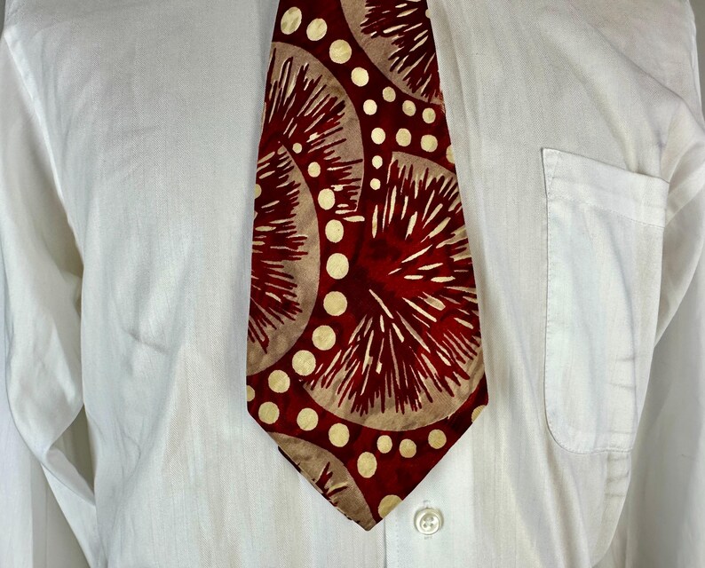 May include: A patterned tie with a red background and beige circular designs with white accents. The tie is worn over a white button-down shirt.