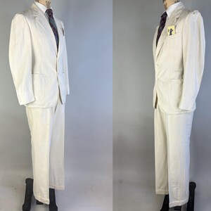 May include: A cream-coloured two-piece suit consisting of a single-breasted blazer with notched lapels, two front pockets, and a chest pocket containing a small yellow tag, paired with matching trousers. A dark tie with a subtle pattern is worn underneath.