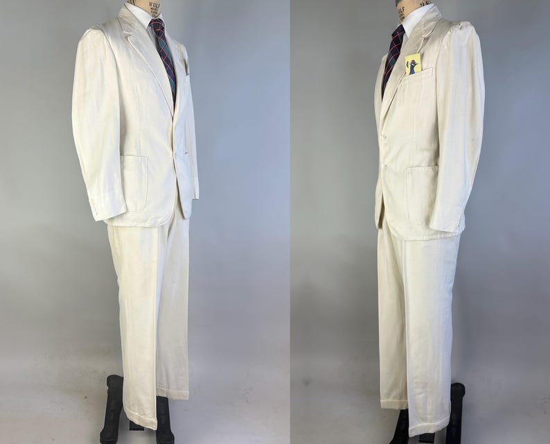 May include: A cream-coloured two-piece suit consisting of a single-breasted blazer with notched lapels, two front pockets, and a chest pocket containing a small yellow tag, paired with matching trousers. A dark tie with a subtle pattern is worn underneath.
