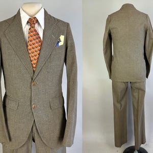 Op de afbeelding: Een beige tweedelig pak bestaande uit een colbert met twee knopen, inkepingen in de revers en klepzakken, gecombineerd met een bijpassende broek. Het colbert wordt gedragen over een wit overhemd met een oranje das met patroon en een pochet.