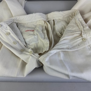 May include: Off-white trousers featuring a button-fly closure with four visible buttons on the waistband and fly. The interior lining is cream-coloured fabric with a rectangular white label containing black printed text and a small red brand tag.