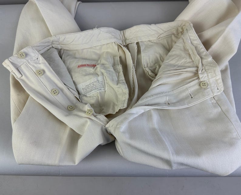 May include: Off-white trousers featuring a button-fly closure with four visible buttons on the waistband and fly. The interior lining is cream-coloured fabric with a rectangular white label containing black printed text and a small red brand tag.