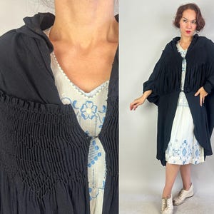 Può includere: Un mantello nero vintage con corpetto a nido d'ape e colletto drappeggiato. Il mantello è aperto sul davanti, rivelando un vestito bianco con ricami floreali blu. L'outfit è completato da scarpe da ginnastica bianche.