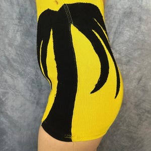 May include: A bright yellow ribbed knit garment featuring a bold, black, abstract palm tree-like graphic pattern that extends vertically along the side. The fabric has a distinct textured, vertical ribbed construction throughout the entire piece.