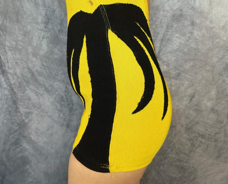 May include: A bright yellow ribbed knit garment featuring a bold, black, abstract palm tree-like graphic pattern that extends vertically along the side. The fabric has a distinct textured, vertical ribbed construction throughout the entire piece.