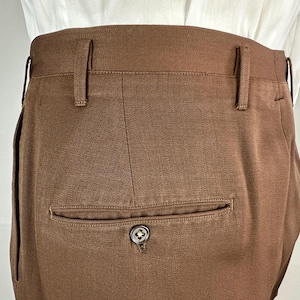 May include: Close-up of brown trousers with belt loops and a single back pocket. The pocket has a button closure with a decorative design. The trousers are made of a textured fabric and appear to be tailored.