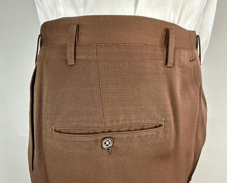 May include: Close-up of brown trousers with belt loops and a single back pocket. The pocket has a button closure with a decorative design. The trousers are made of a textured fabric and appear to be tailored.