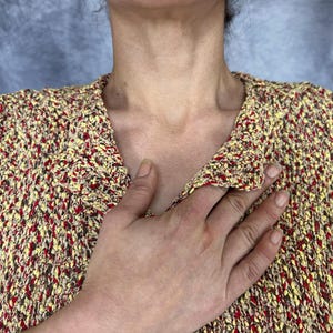 May include: A textured, open-weave sweater in a mix of yellow, red, and brown. The sweater has a V-neck and a loose fit. The image shows the upper body and part of a hand.