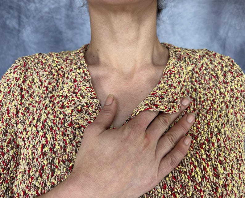 May include: A textured, open-weave sweater in a mix of yellow, red, and brown. The sweater has a V-neck and a loose fit. The image shows the upper body and part of a hand.