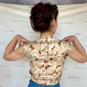 May include: A cream-colored, short-sleeved top with a butterfly and foliage pattern in shades of brown and orange. The top has a high collar and a button-up back. The fabric appears to be a satin or silk material.