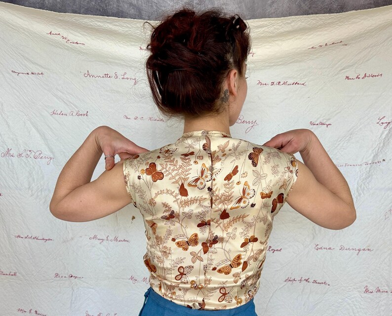 May include: A cream-colored, short-sleeved top with a butterfly and foliage pattern in shades of brown and orange. The top has a high collar and a button-up back. The fabric appears to be a satin or silk material.