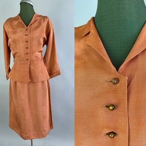 May include: A peach-colored two-piece suit with a long-sleeved jacket and a skirt. The jacket has a collar and a button-up front. The skirt is knee-length and has a straight silhouette.