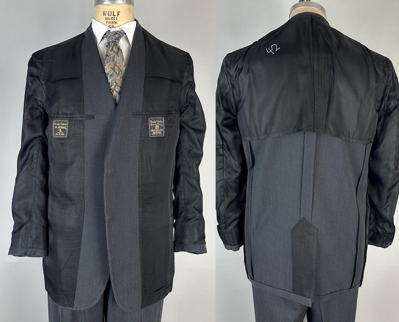 May include: A charcoal gray blazer shown from the front and back, featuring a partial black lining, a shawl collar, and a patterned necktie. The interior lining displays two rectangular labels with text and the number 42 written on the back fabric.