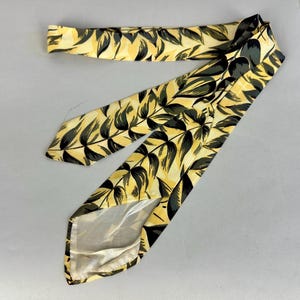 May include: A yellow and black patterned tie with a leaf design. The tie is made of silk and has a pointed tip.