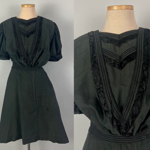 May include: A vintage black dress with short, puffed sleeves and a fitted waist. The neckline and bodice feature decorative black velvet trim. The dress has a flared skirt and is displayed on a mannequin.