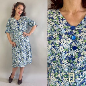Könnte beinhalten: Vintage Kleid mit floralem Muster, V-Ausschnitt und kurzen Ärmeln. Das Kleid hat blaue Knöpfe und einen passenden Gürtel mit rechteckiger Schnalle. Der Stoff zeigt ein Muster aus weißen, blauen und grünen Blumen.