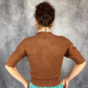Pu&ograve; includere: Un maglione marrone a maniche corte con una trama a coste. Il maglione ha uno scollo alto con un colletto decorativo e una cerniera sul retro. Le maniche sono lunghe fino al gomito.