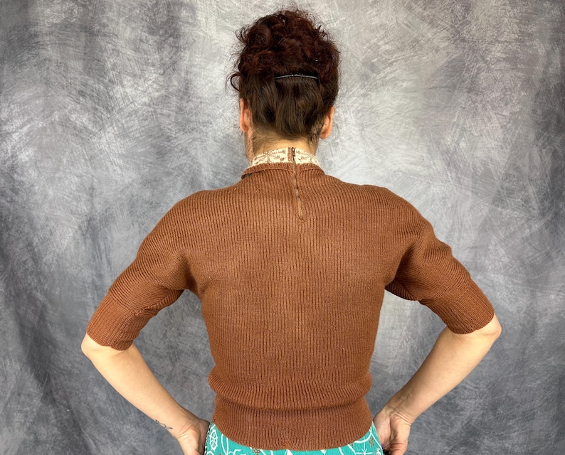 Pu&ograve; includere: Un maglione marrone a maniche corte con una trama a coste. Il maglione ha uno scollo alto con un colletto decorativo e una cerniera sul retro. Le maniche sono lunghe fino al gomito.