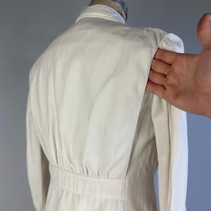 May include: An off-white jacket featuring a structured collar, long sleeves, and a fitted waistline. The back panel displays vertical pleats that extend from the shoulder down to the waist seam, creating a tailored silhouette with a clean finish.